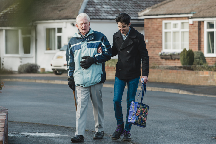 Teenage boy is walking back from the shop with an older gentleman. The boy is carrying the shopping bag and they are linking arms.
