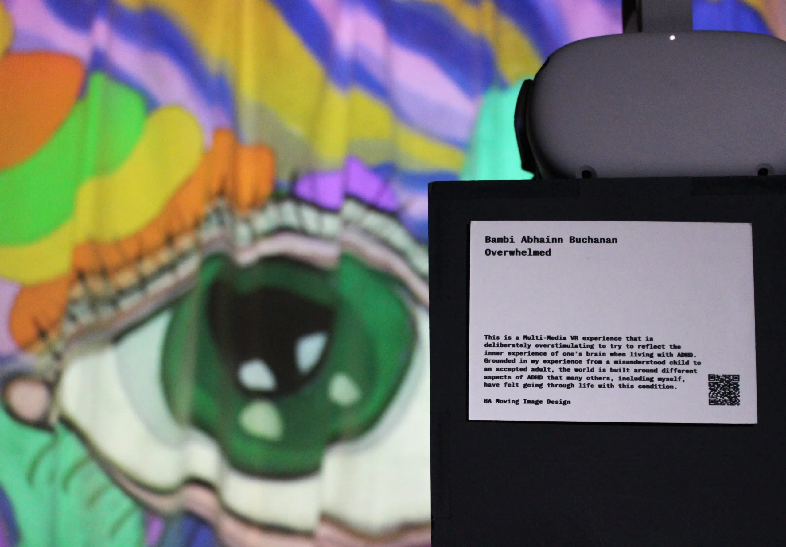An art installation on display, featuring a colourful illustration of an eye being projected on to a large cloth. On the back of the projector, there is a description of the installation, with the artist's name and the name of the art installation.